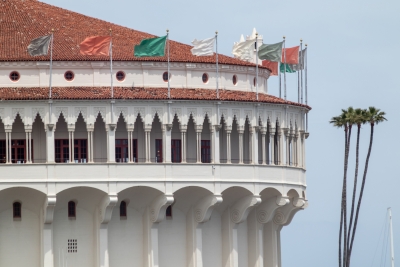 Close-up of the Catalina Casino's arched colonnade and red-tile roof, colorful flags flying above against a clear sky.